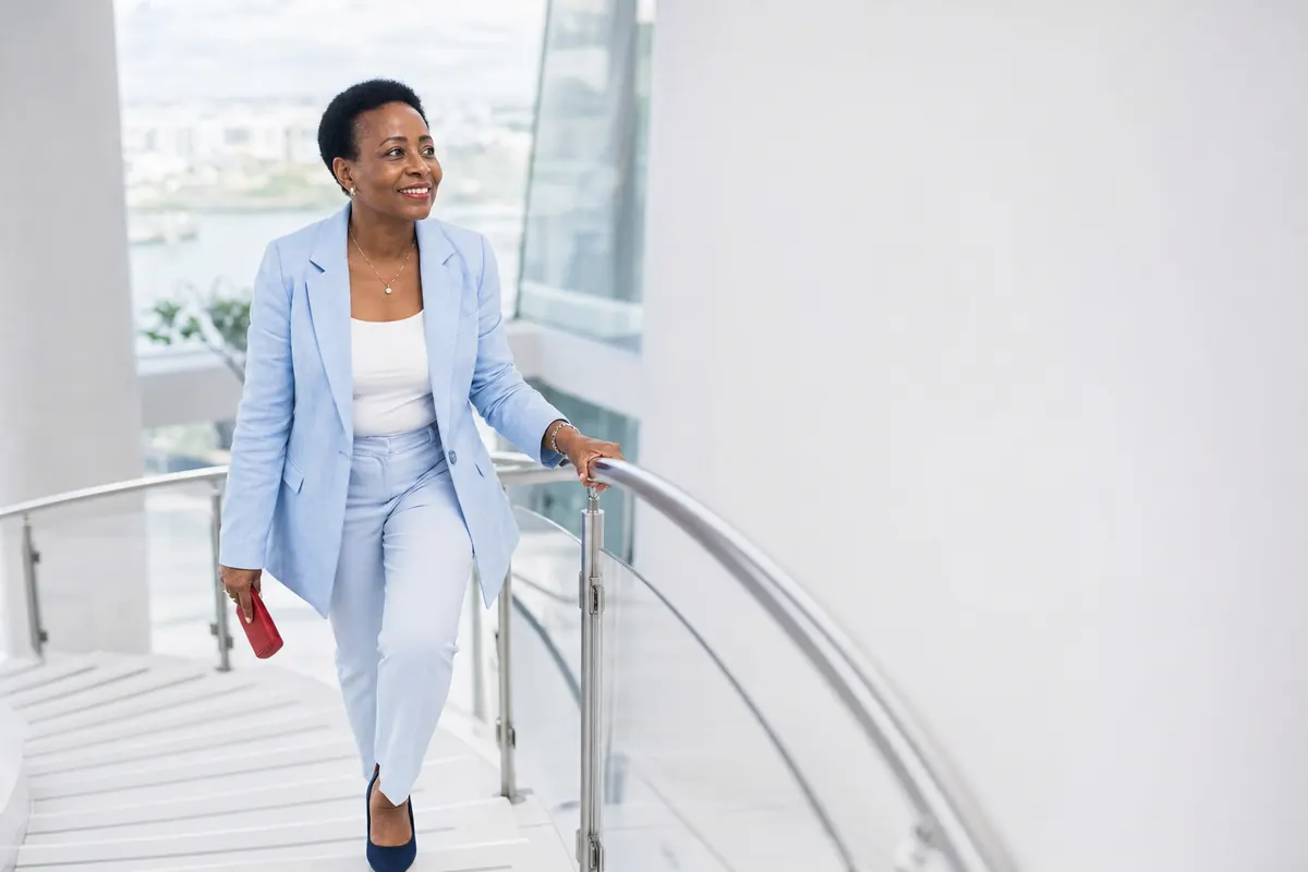 Smiling Nigerian woman in light blue blazer climbing bright white staircase with ease